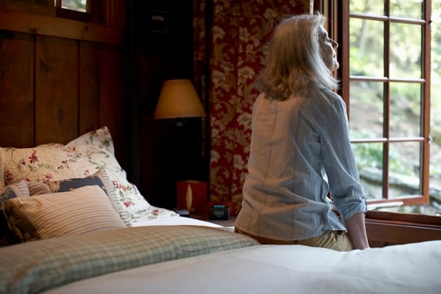 A middle-aged woman sitting on a bed, seen from behind, gazing out a window, reflecting the quiet inner weight of overfunctioning in relationships.