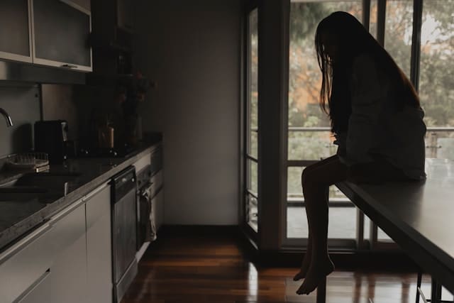 why am I so mean to myself — a woman sitting alone in a kitchen, head bowed, looking down